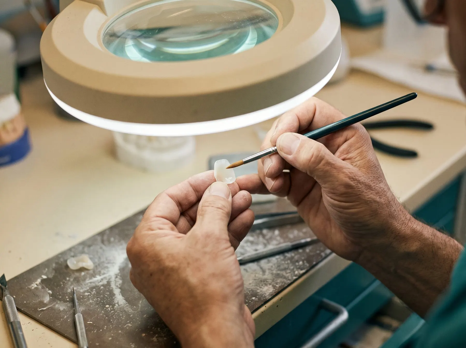 Ceramist shaping a porcelain veneer by hand under a magnifying lamp